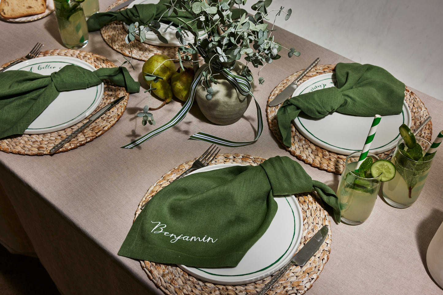 Earthy table setting with green linen napkin embroidered Benjamin, paired with woven placemats and fresh citrus drinks