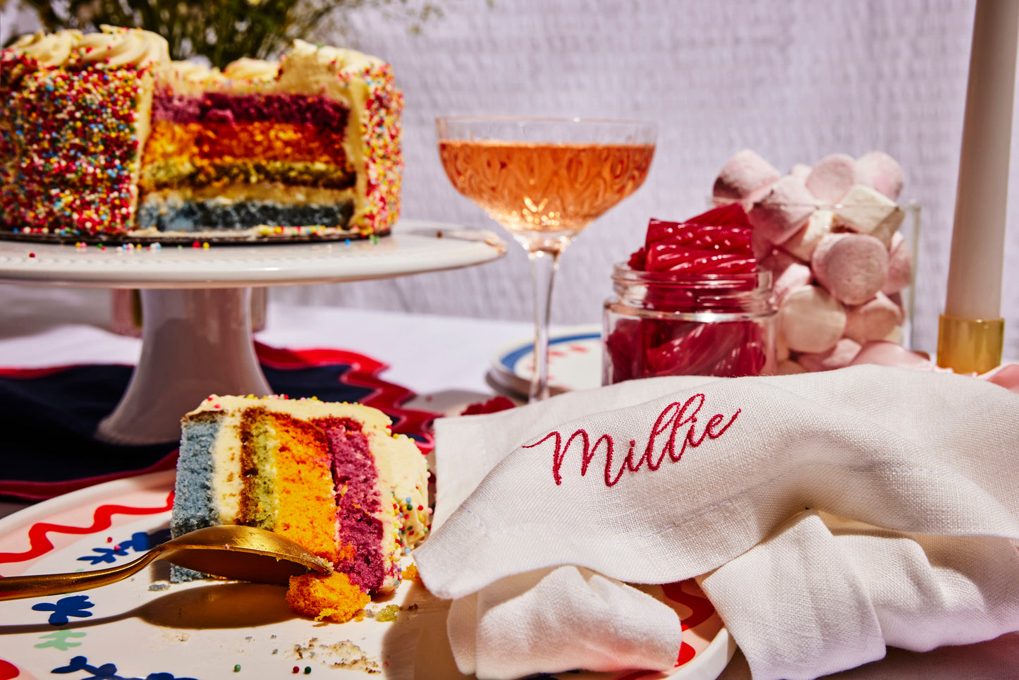 Custom white linen napkin with Millie embroidery, styled beside rainbow cake, pink sweets, and sparkling rosé