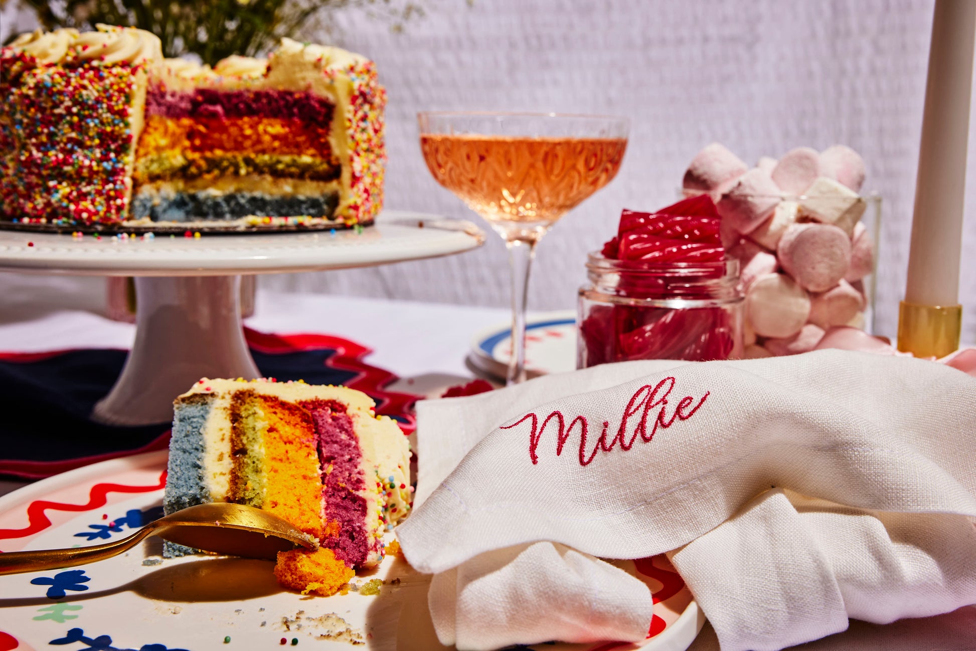 Custom white linen napkin with Millie embroidery, styled beside rainbow cake, pink sweets, and sparkling rosé