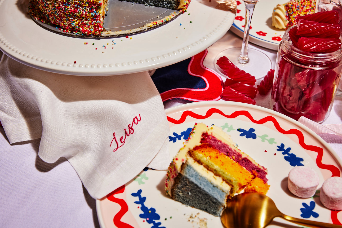 White linen napkin with pink embroidered name Leisa beside rainbow birthday sprinkle cake and colourful tableware