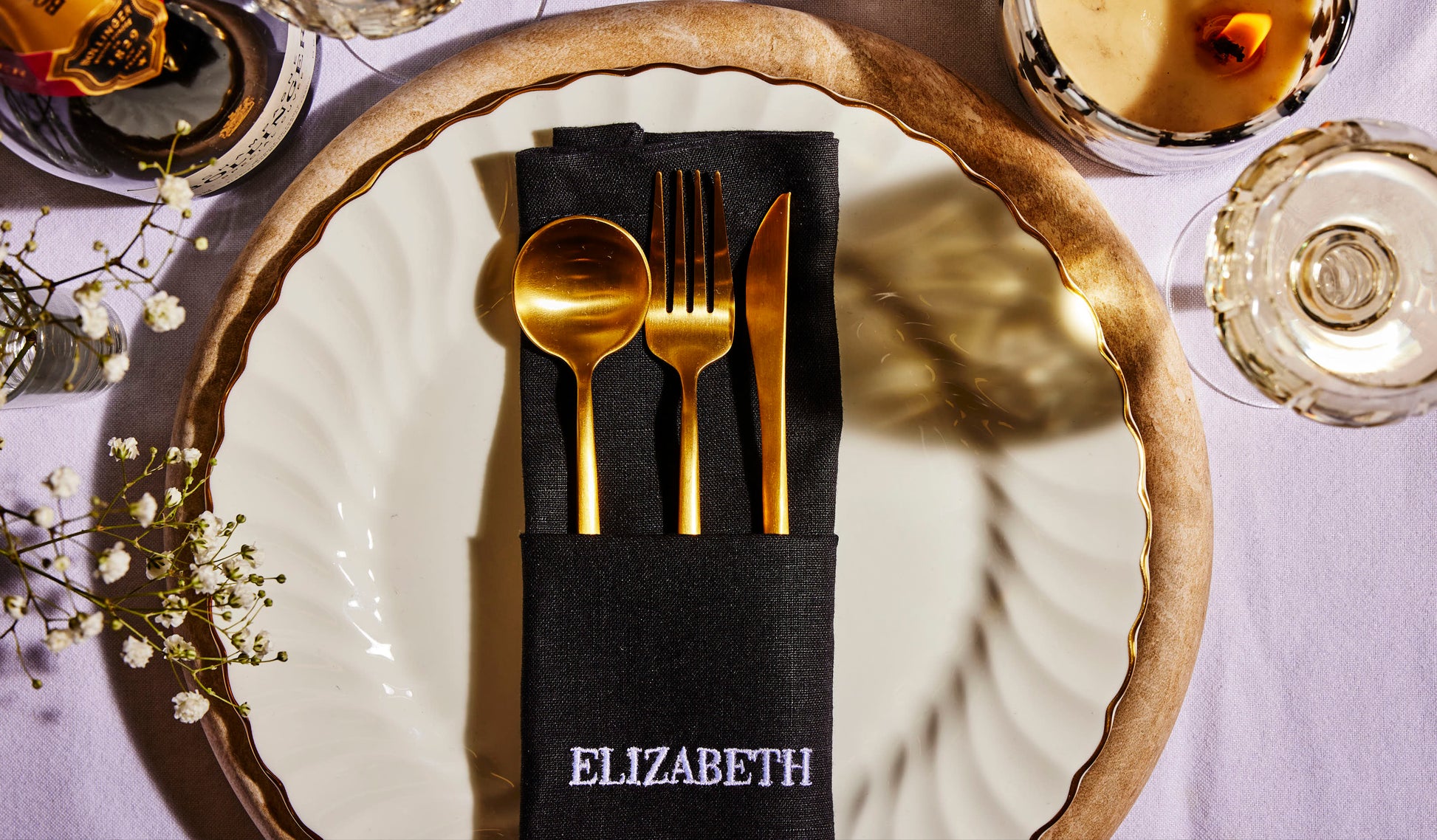 Black linen napkin embroidered Elizabeth in white, styled with gold cutlery on a formal table setting with champagne.