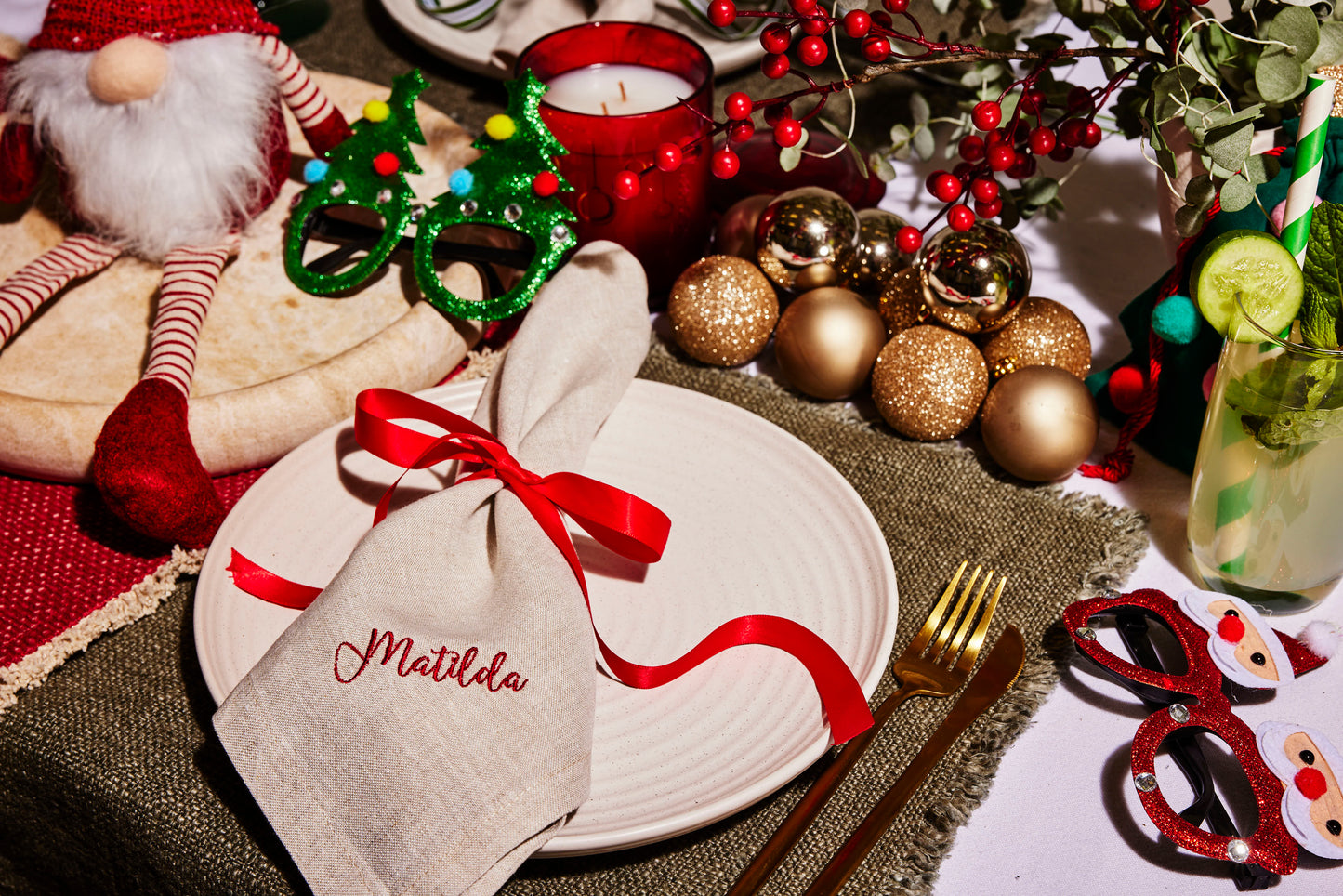 Beige linen napkin embroidered Matilda with red ribbon, styled on a festive Christmas table with gold baubles and novelty glasses