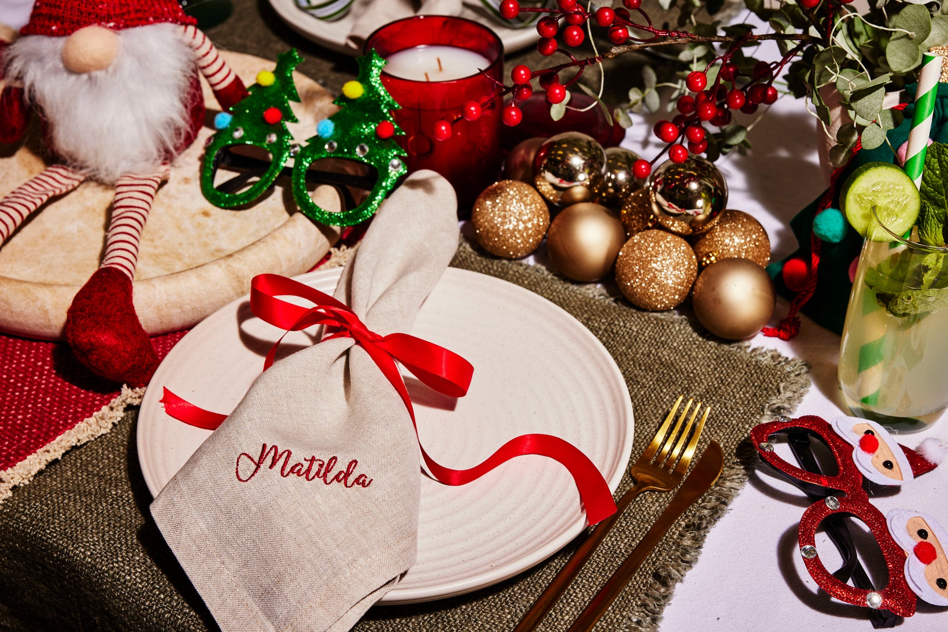 Beige linen napkin embroidered Matilda with red ribbon, styled on a festive Christmas table with gold baubles and novelty glasses