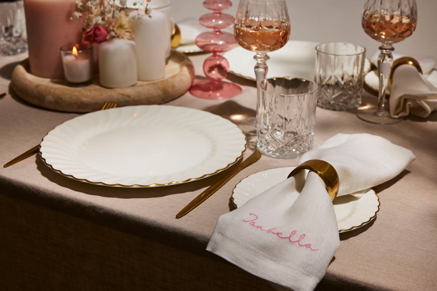Personalised white linen napkin embroidered Isabella in pink, styled with gold ring and soft candlelit table setting.