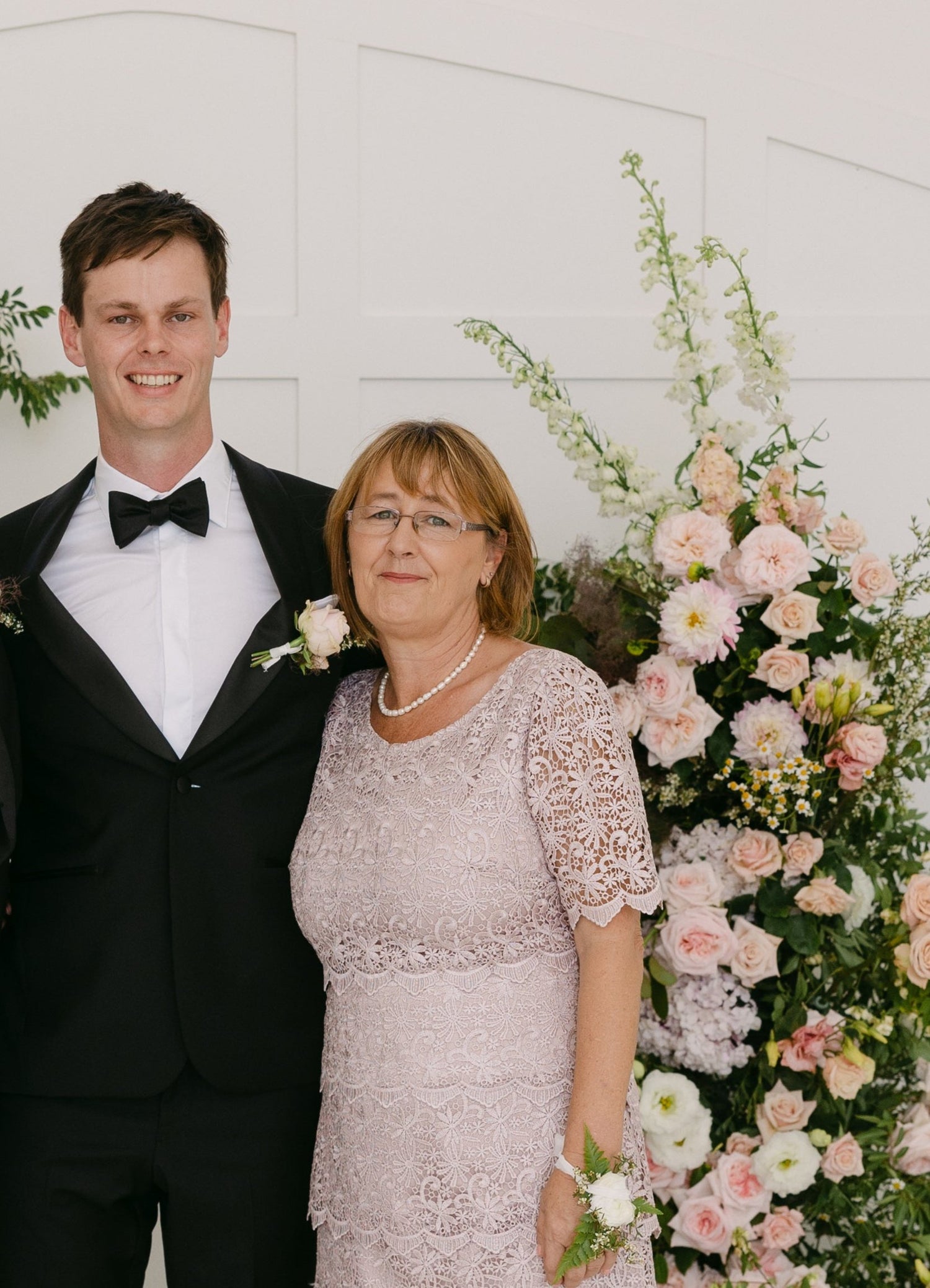 Mother and son smiling together in formal wedding attire, standing beside a floral arrangement of blush and ivory flowers.