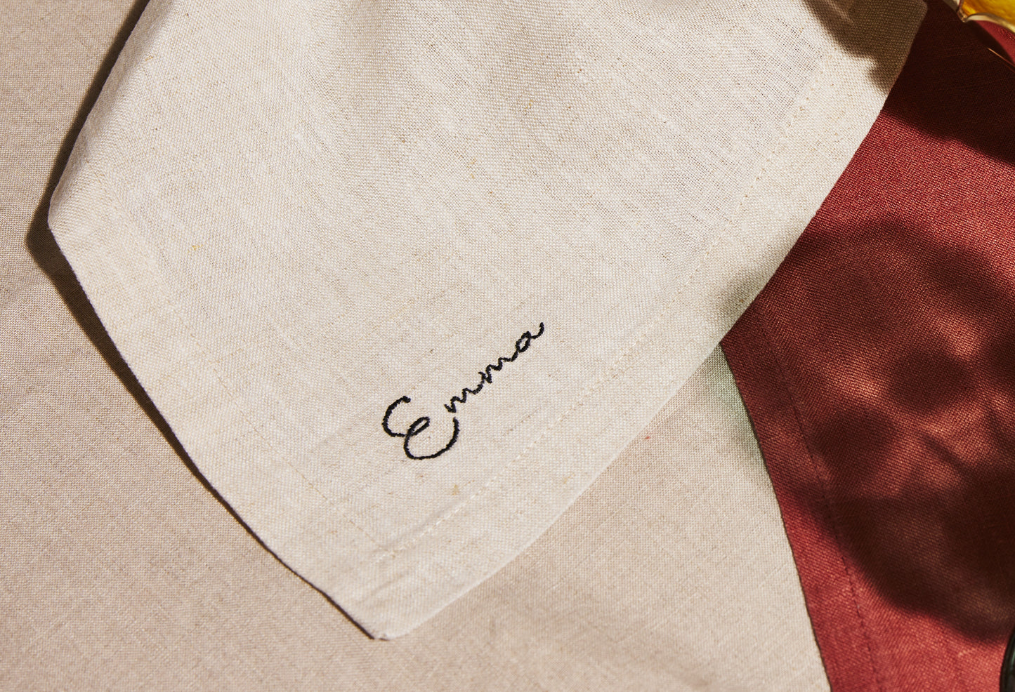 Natural linen napkin with black embroidered name Emma, styled on warm-toned fabric backdrop