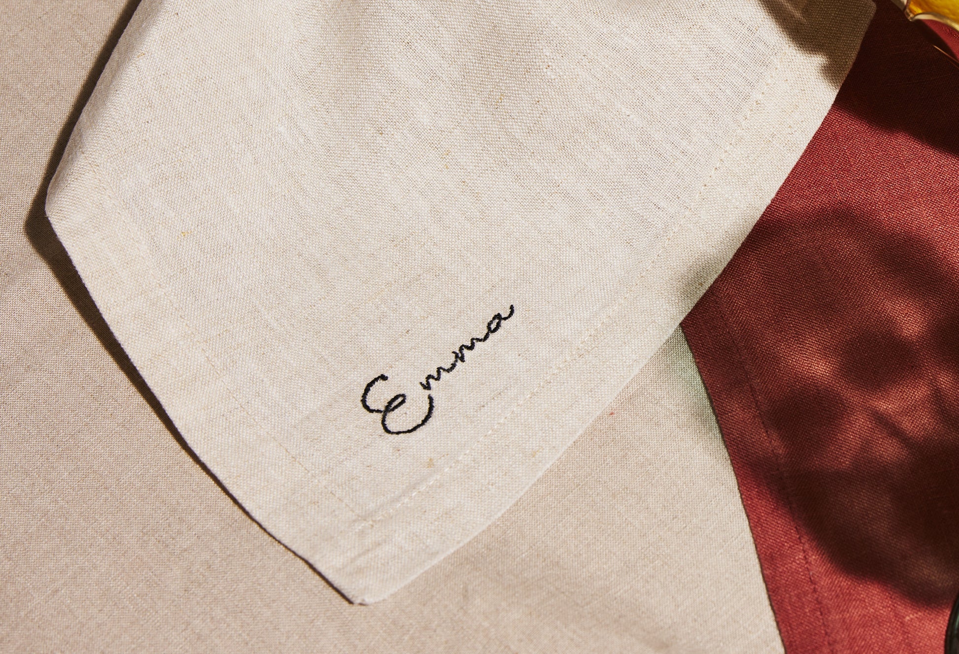 Natural linen napkin with black embroidered name Emma, styled on warm-toned fabric backdrop