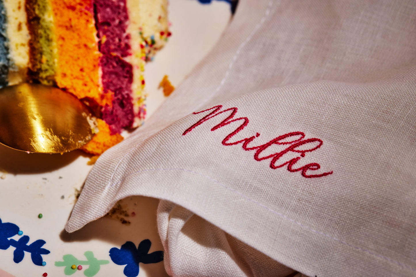 Personalised linen napkin with red embroidered name Millie beside colourful layered cake and gold spoon