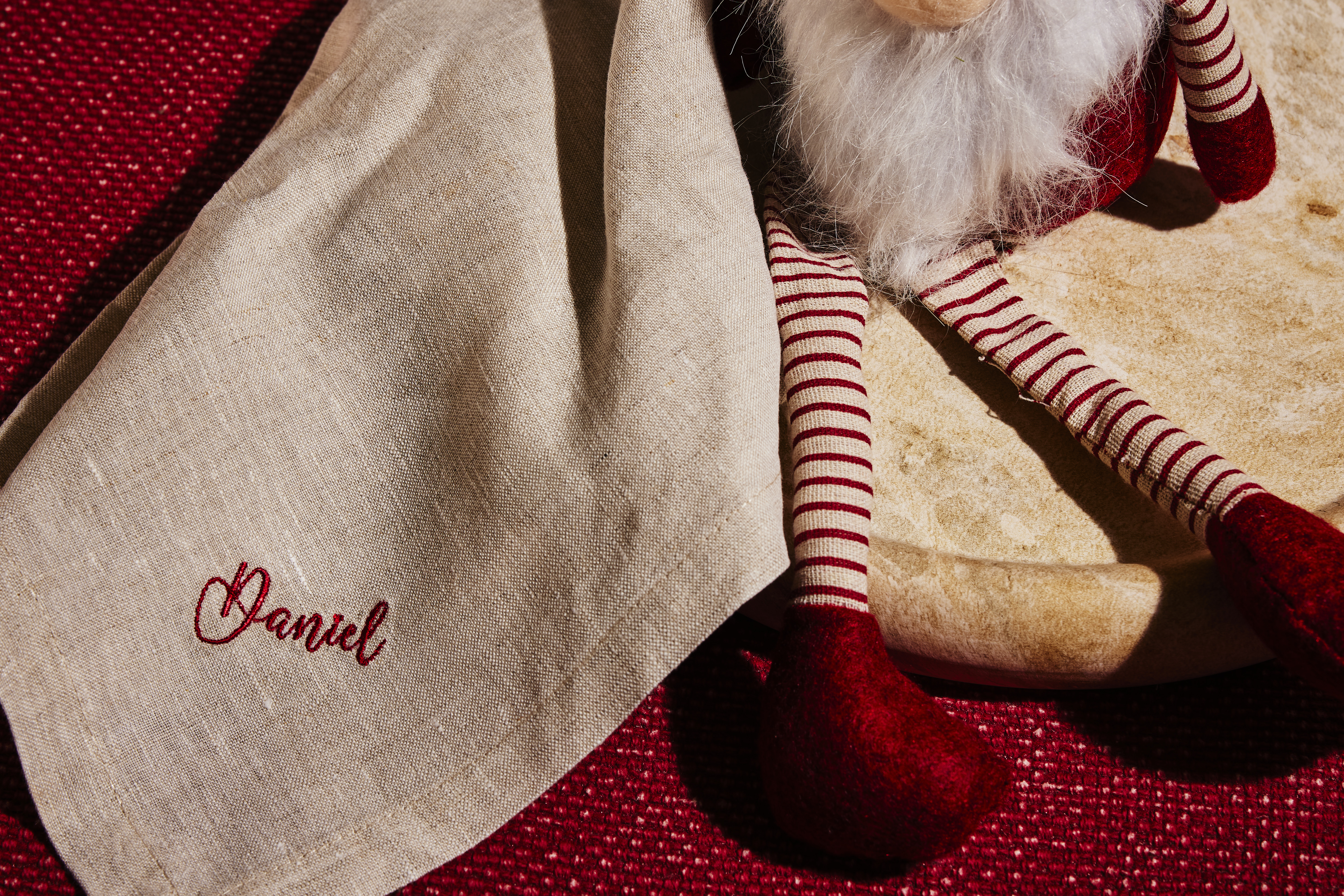Natural linen napkin embroidered "Daniel" in red script on a red textured background beside striped red-and-white toy legs