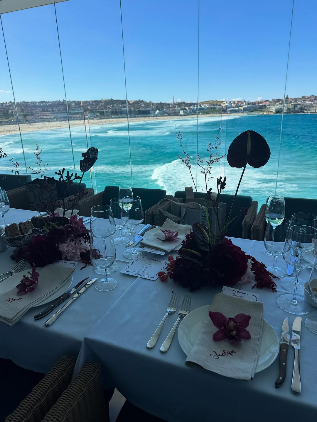 Personalised off-white linen napkins with embroidery at a seaside table setting, dark red orchids and glassware against a turquoise ocean view