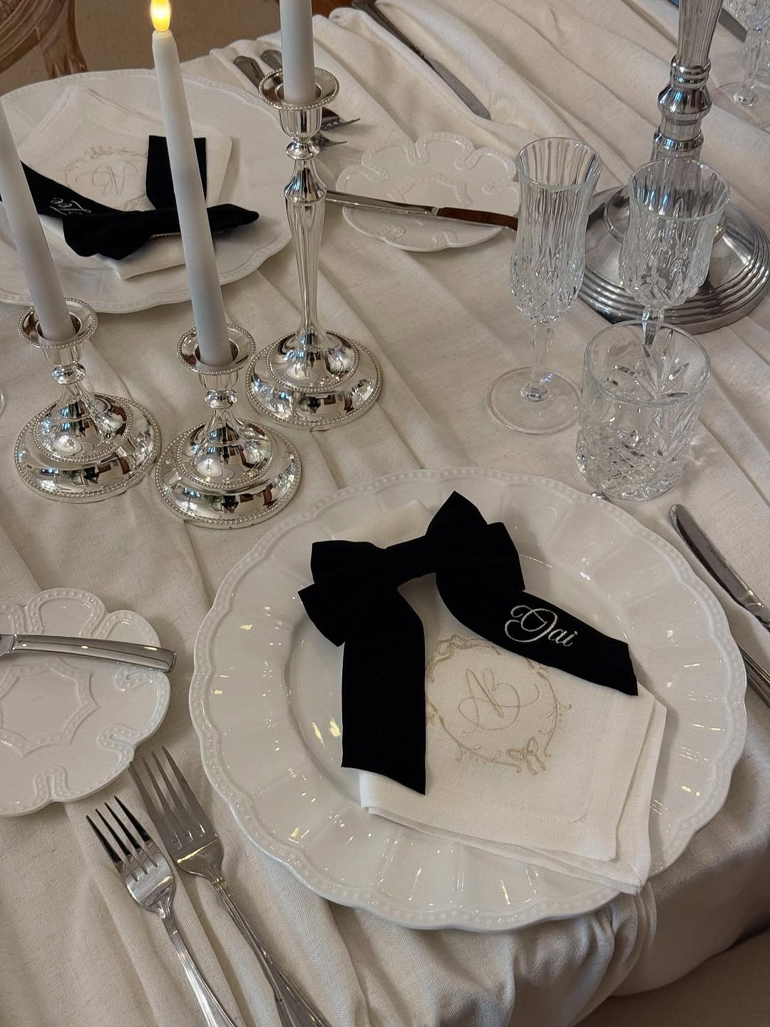 Cream linen tablecloth with a white embroidered monogram napkin on a scalloped plate, topped by a black velvet bow and silver candlesticks