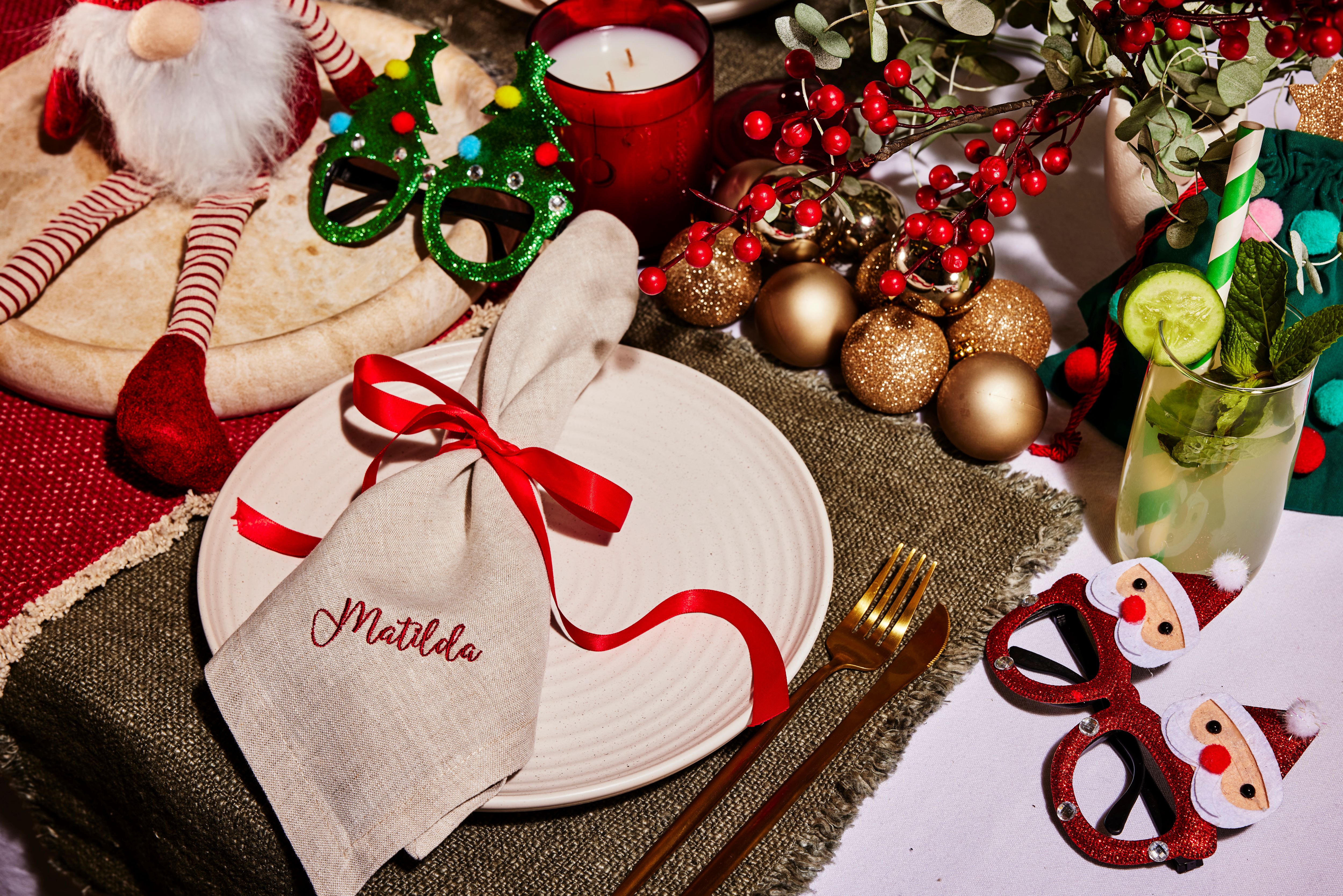 Beige linen napkin embroidered Matilda in red, tied with a red ribbon on a plate amid gold baubles, red berries and festive props.