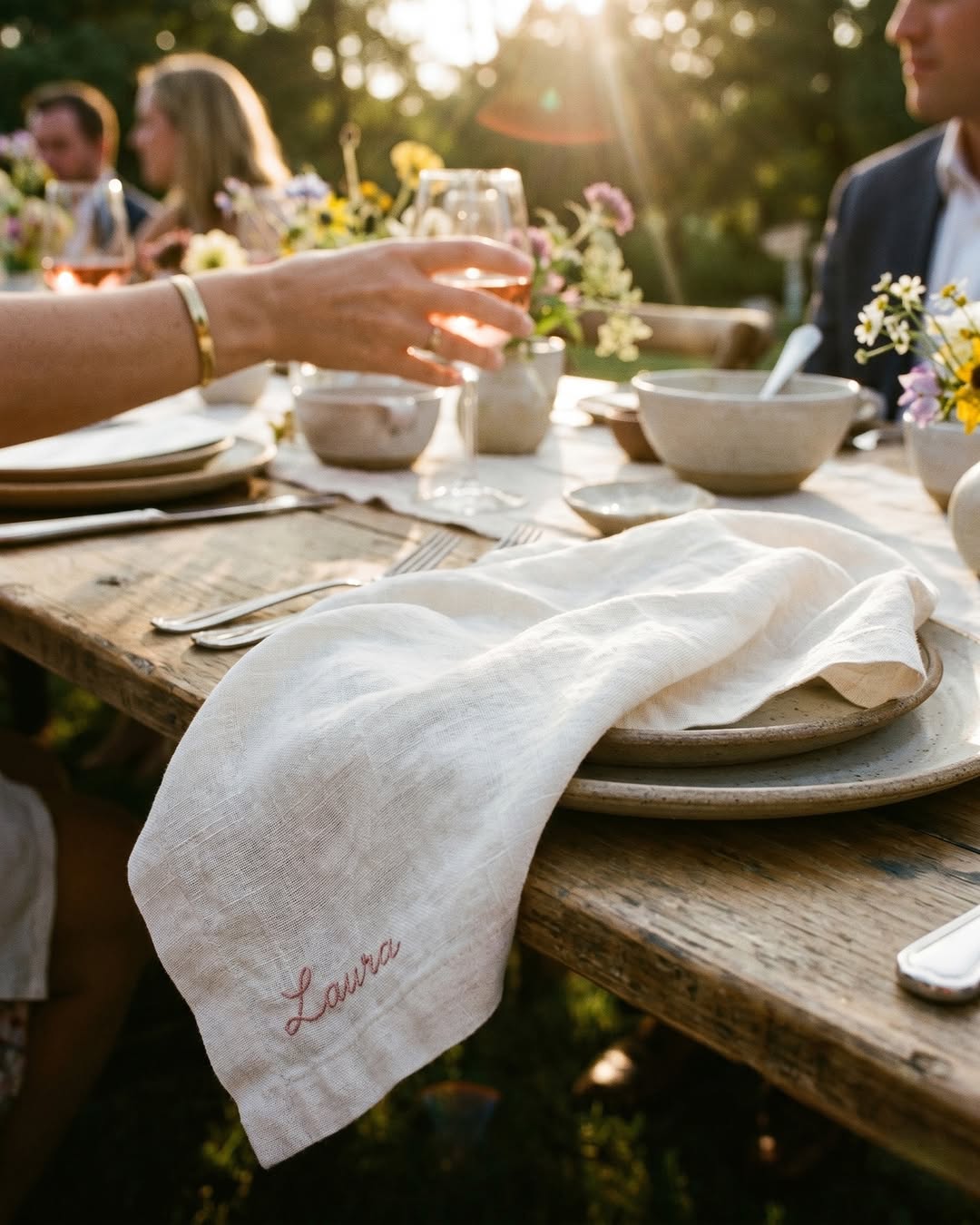 Soft white linen napkin embroidered "Laura" in pink on a rustic wooden table set with cream stoneware, wildflowers and warm evening light.