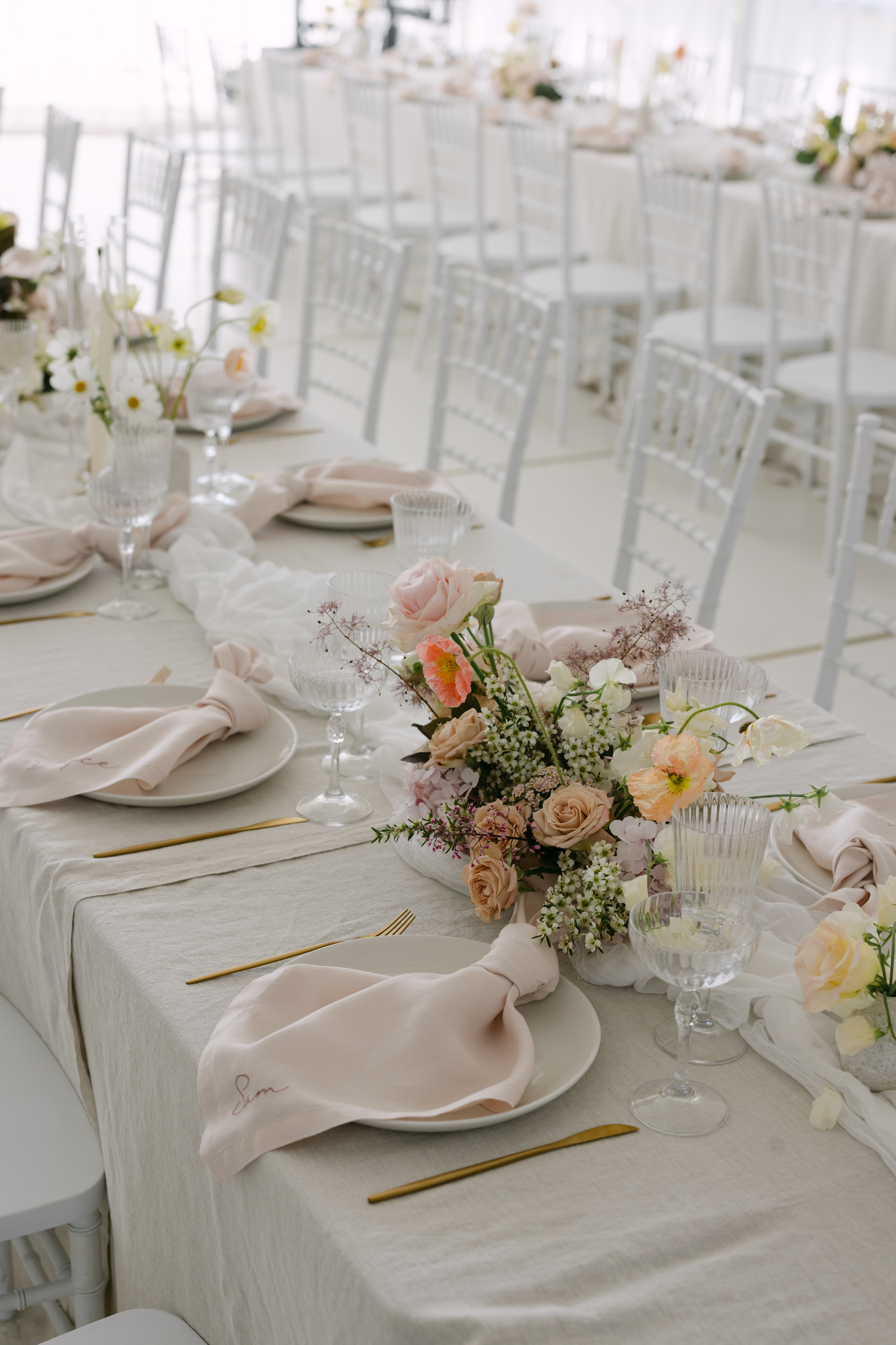 Cream linen tablecloth with pale blush embroidered napkins, gold cutlery, crystal glassware and mixed floral centrepieces