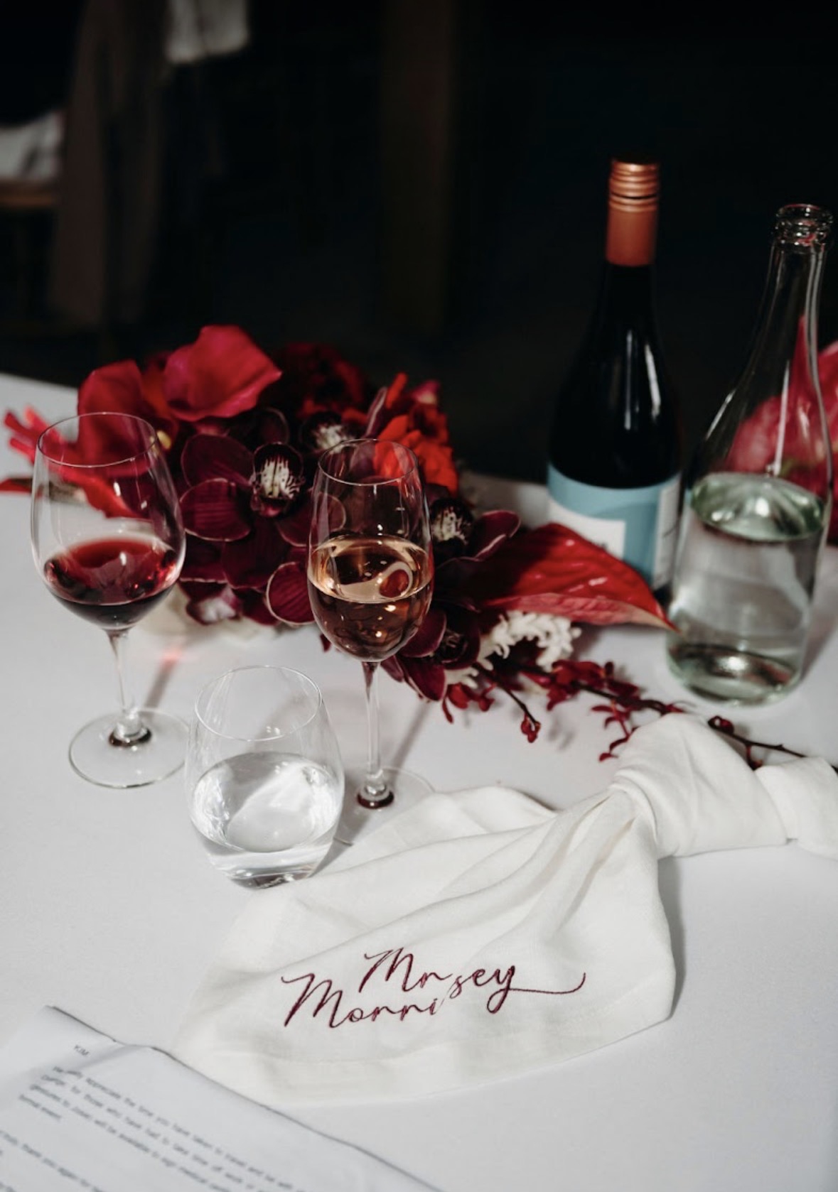 White knotted linen napkin with burgundy embroidery on a white tablecloth beside red floral centerpiece and wine glasses.