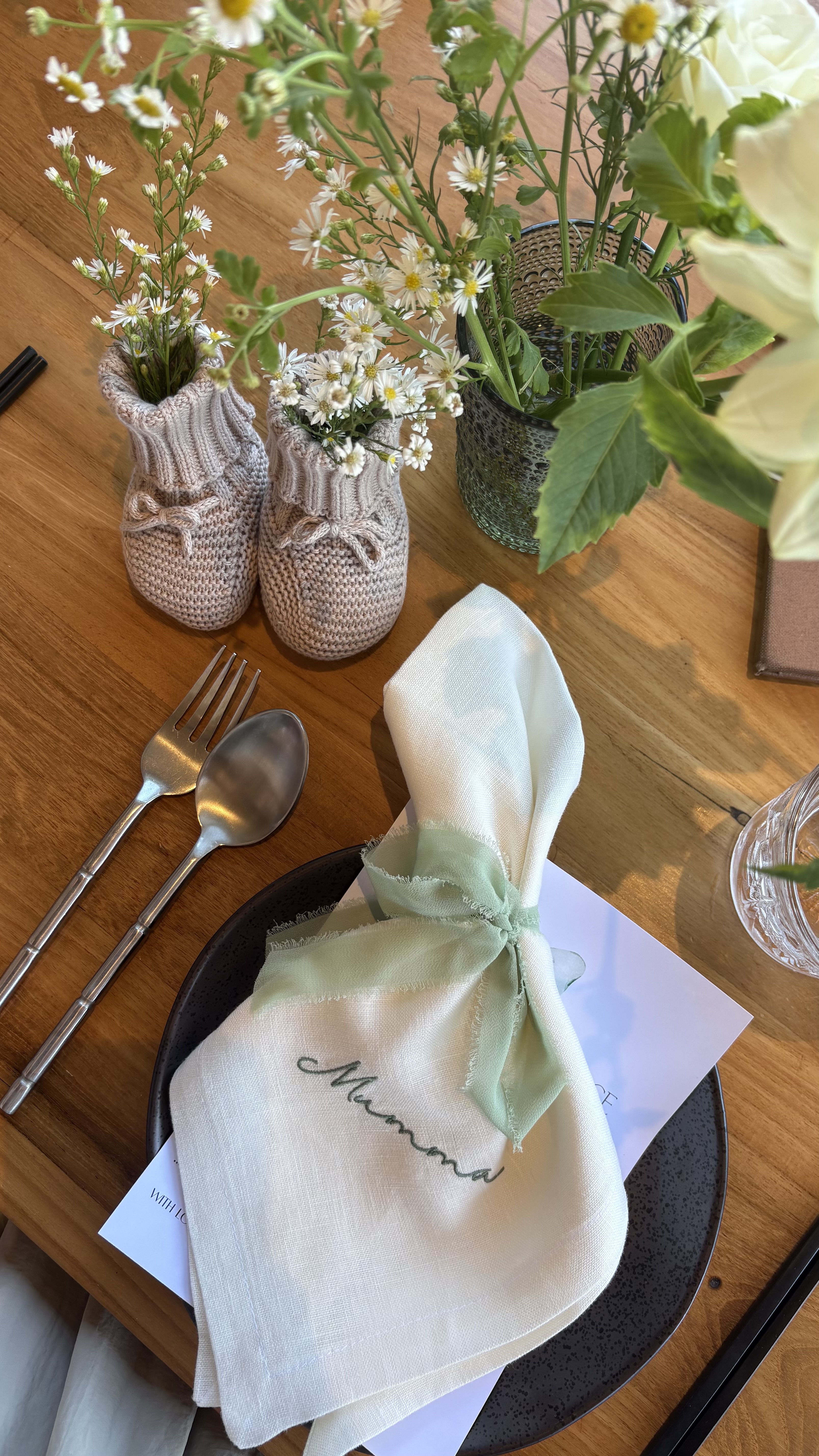 Cream linen napkin with green embroidered name and sage chiffon ribbon on a black speckled plate beside cutlery and wildflowers