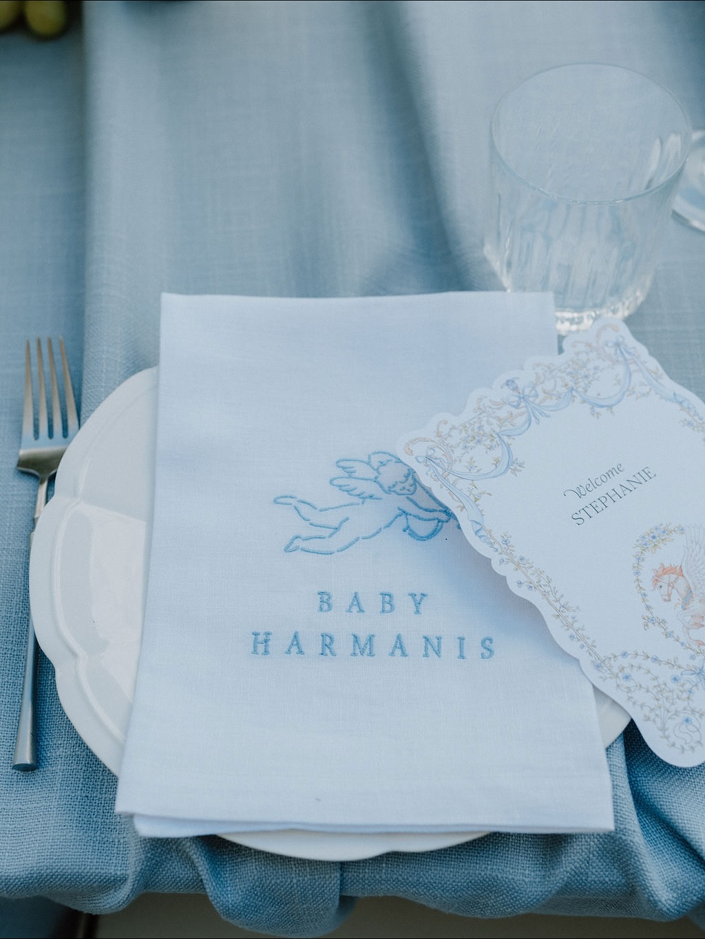 White linen napkin with blue cherub embroidery and "BABY HARMANIS" lettering on a white plate over a textured blue tablecloth