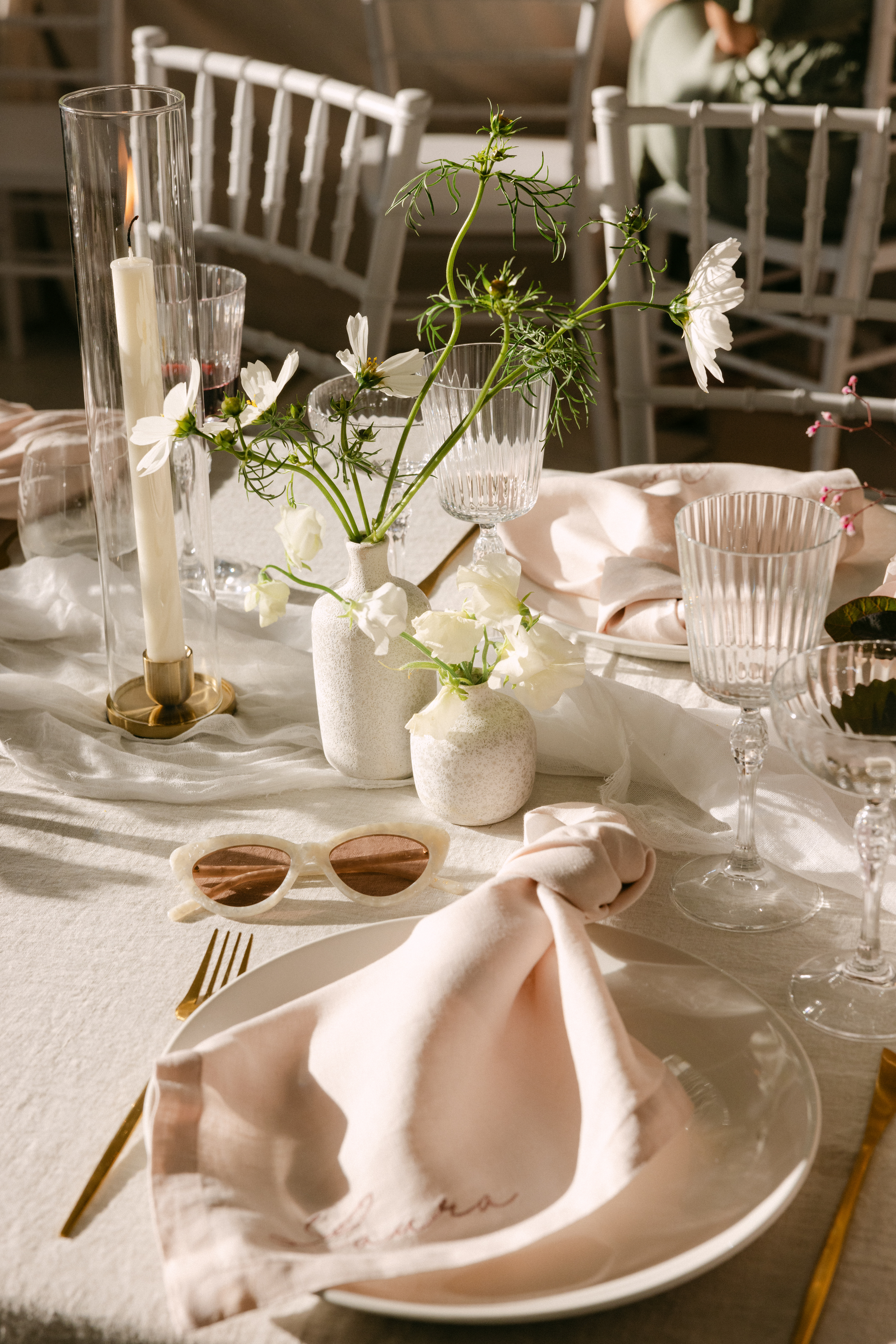 Blush linen napkin with embroidered corner on white plate, gold cutlery, ribbed glassware and white florals in textured vases