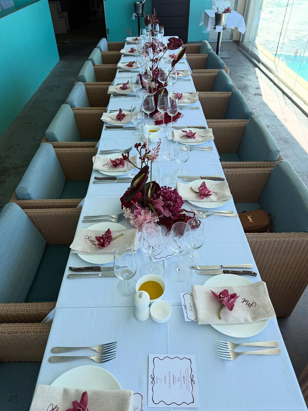 Long white-tablecloth dining table with natural linen embroidered napkins, burgundy orchids and matching floral runners by a pool