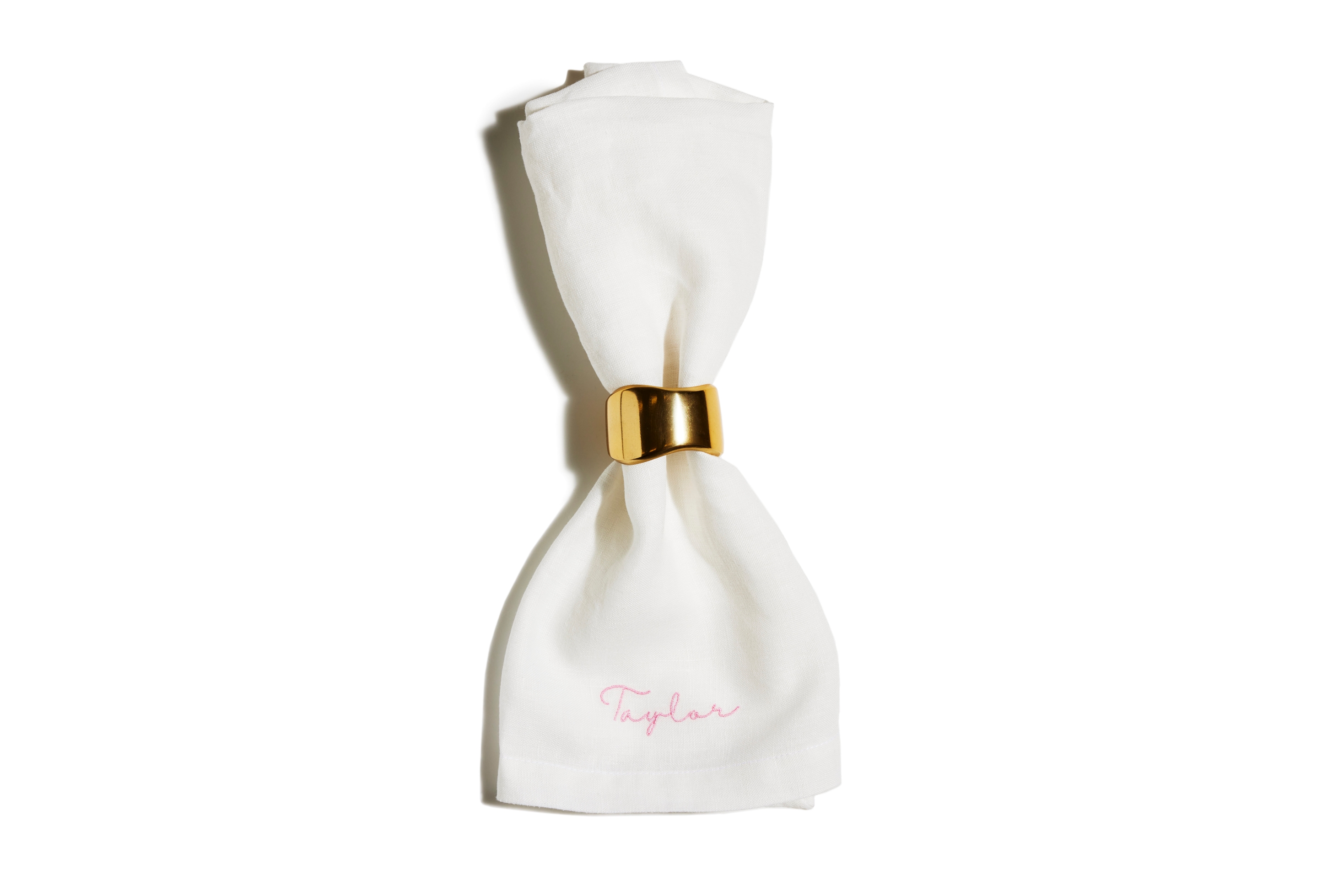 White linen napkin folded through a gold napkin ring with pink script "Taylor" embroidered at the hem