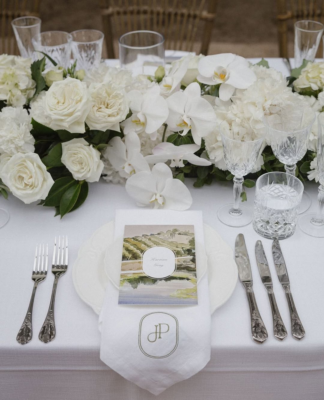 White linen napkin with gold embroidered monogram tucked under a plate amid white roses and orchids, crystal glassware and silver cutlery