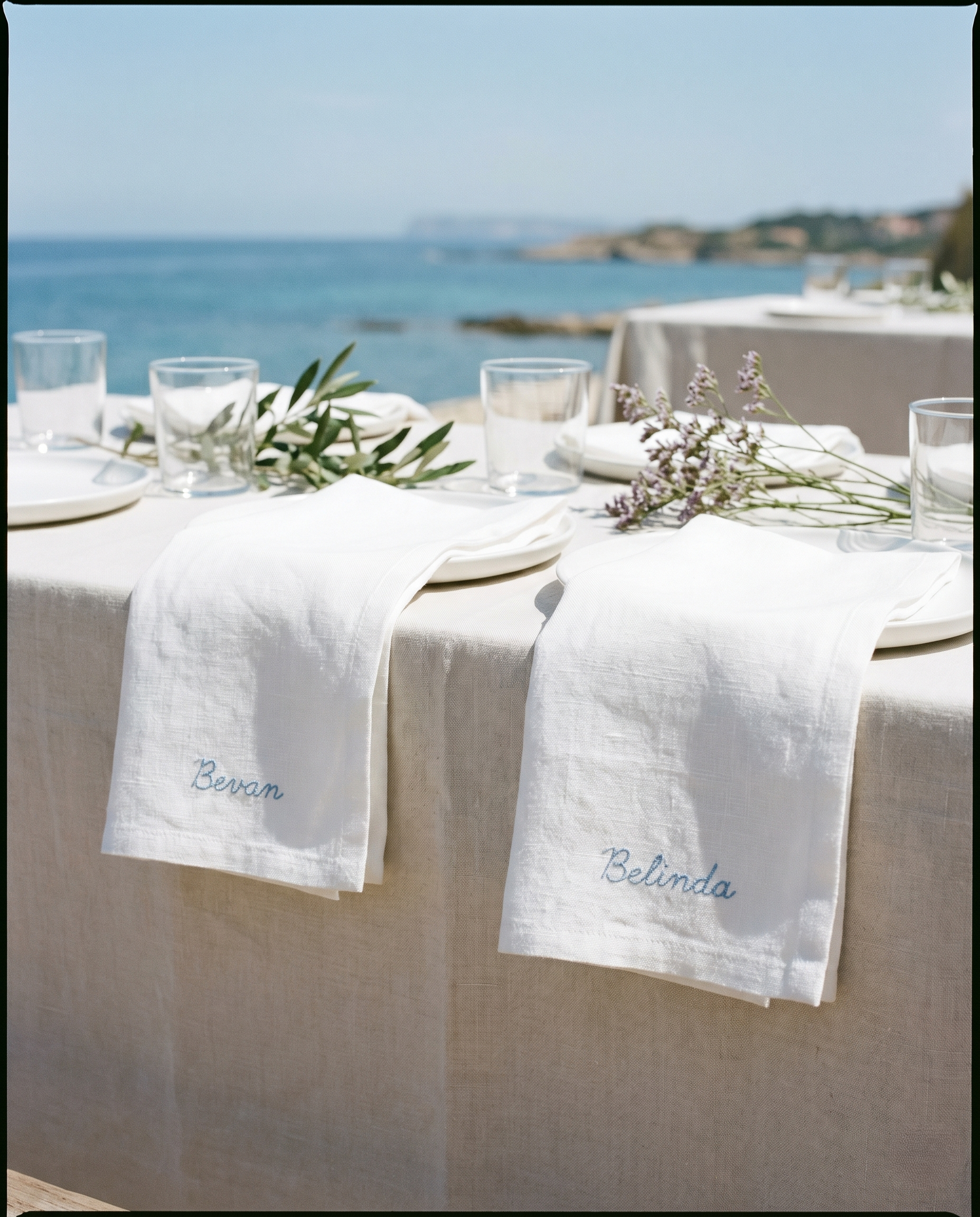 White linen napkins embroidered "Bevan" and "Belinda" in blue drape over plates on a seaside table with glassware and sprigs