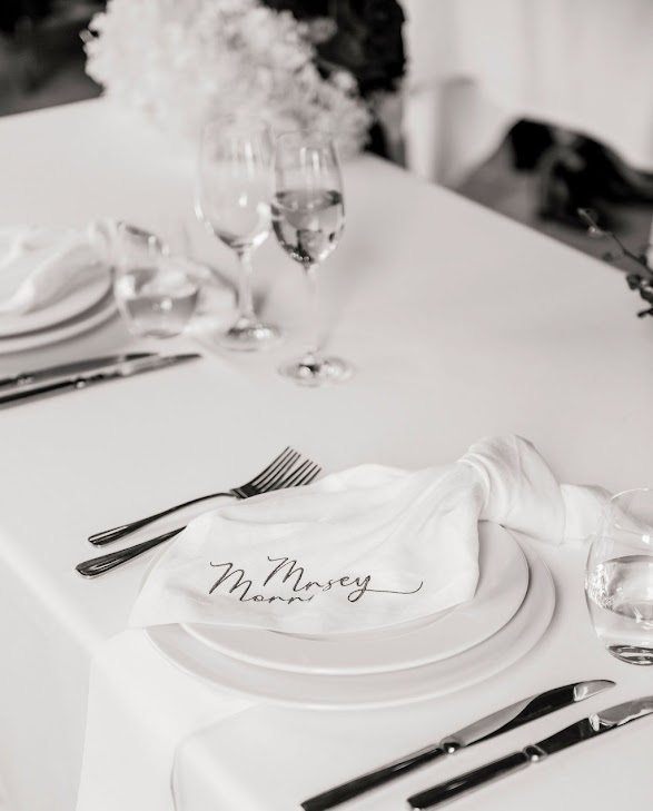 White linen napkin with dark script embroidery styled on stacked white plates at an elegant table setting with glassware