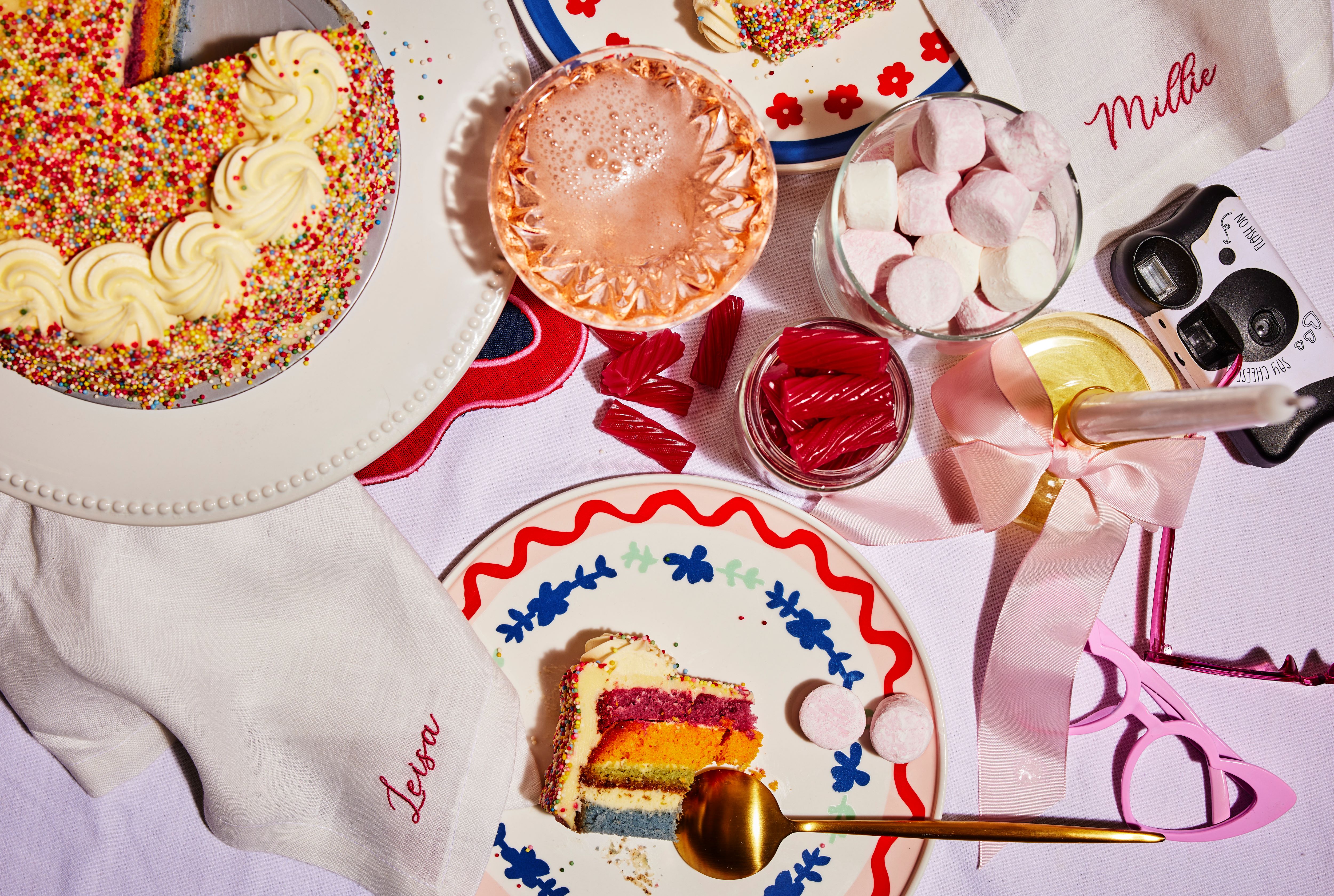 White linen napkin embroidered in red beside a colorful layered cake slice, candies and pink glassware on a styled table