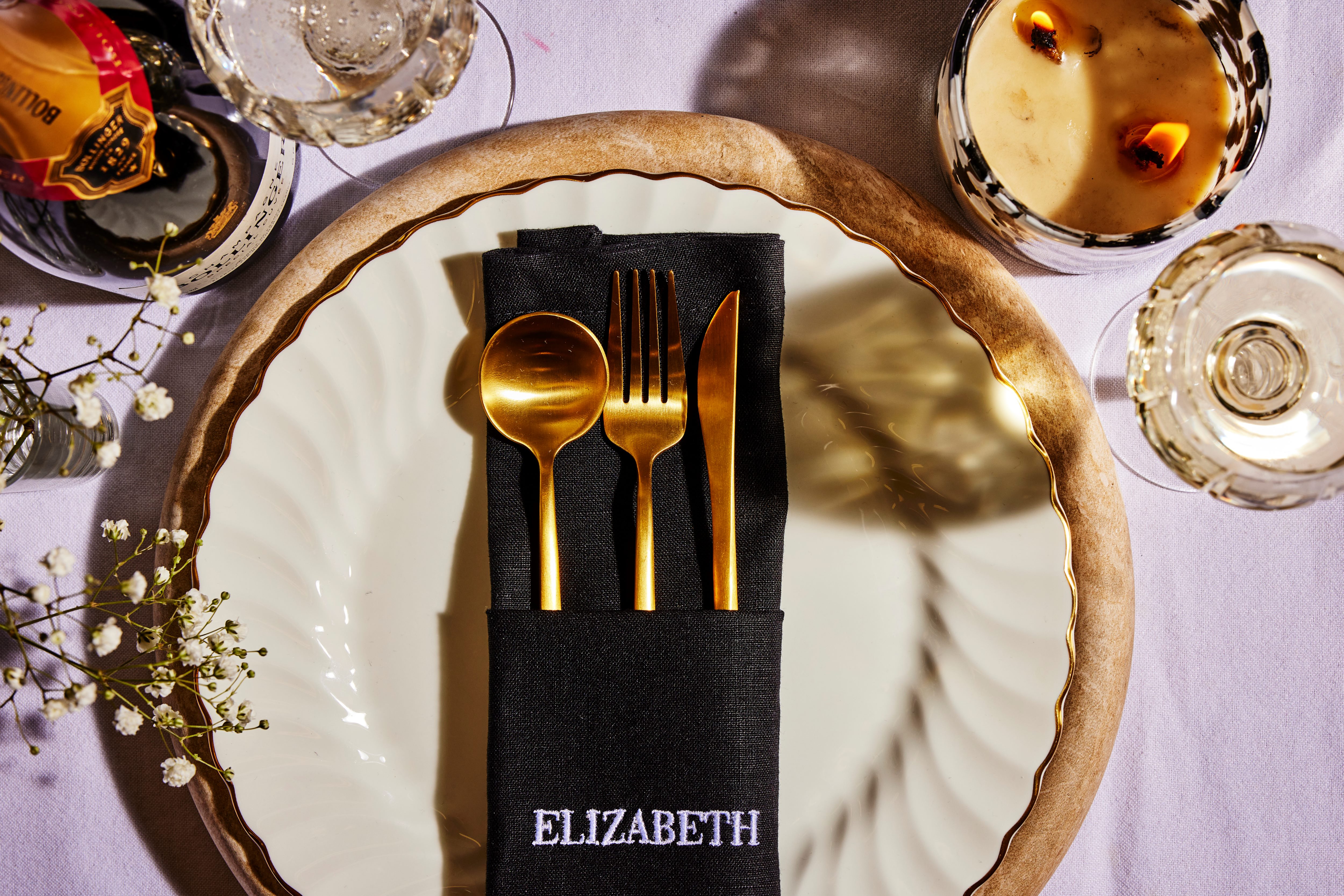 Black linen pocket napkin embroidered "ELIZABETH" holding gold cutlery on cream scalloped plate with wooden charger and lilac tablecloth