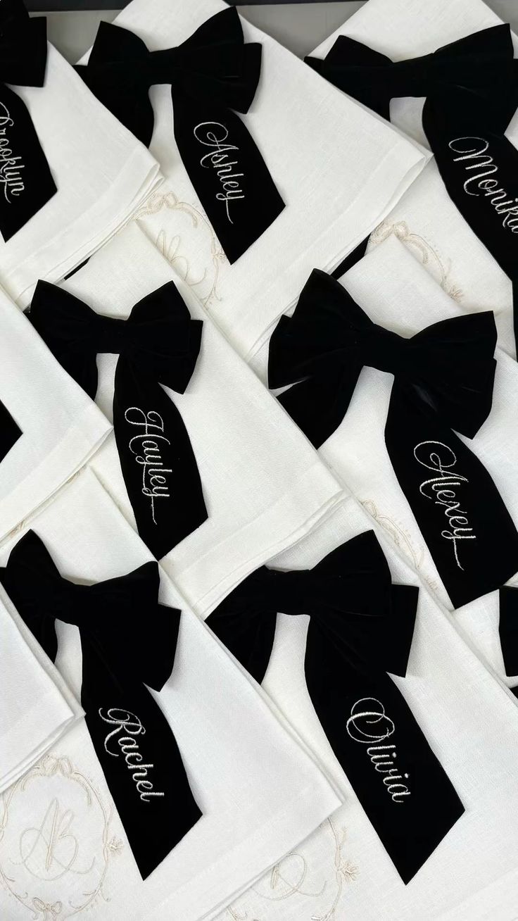 Crisp white linen napkins arranged overlapping, each tied with a black velvet bow and personalised white-script embroidered tags.