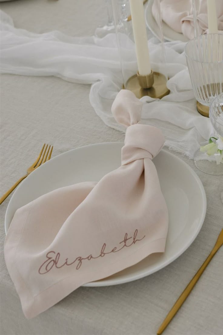 Pale blush linen napkin with embroidered script name draped on a white plate over a natural linen tablecloth with gold cutlery
