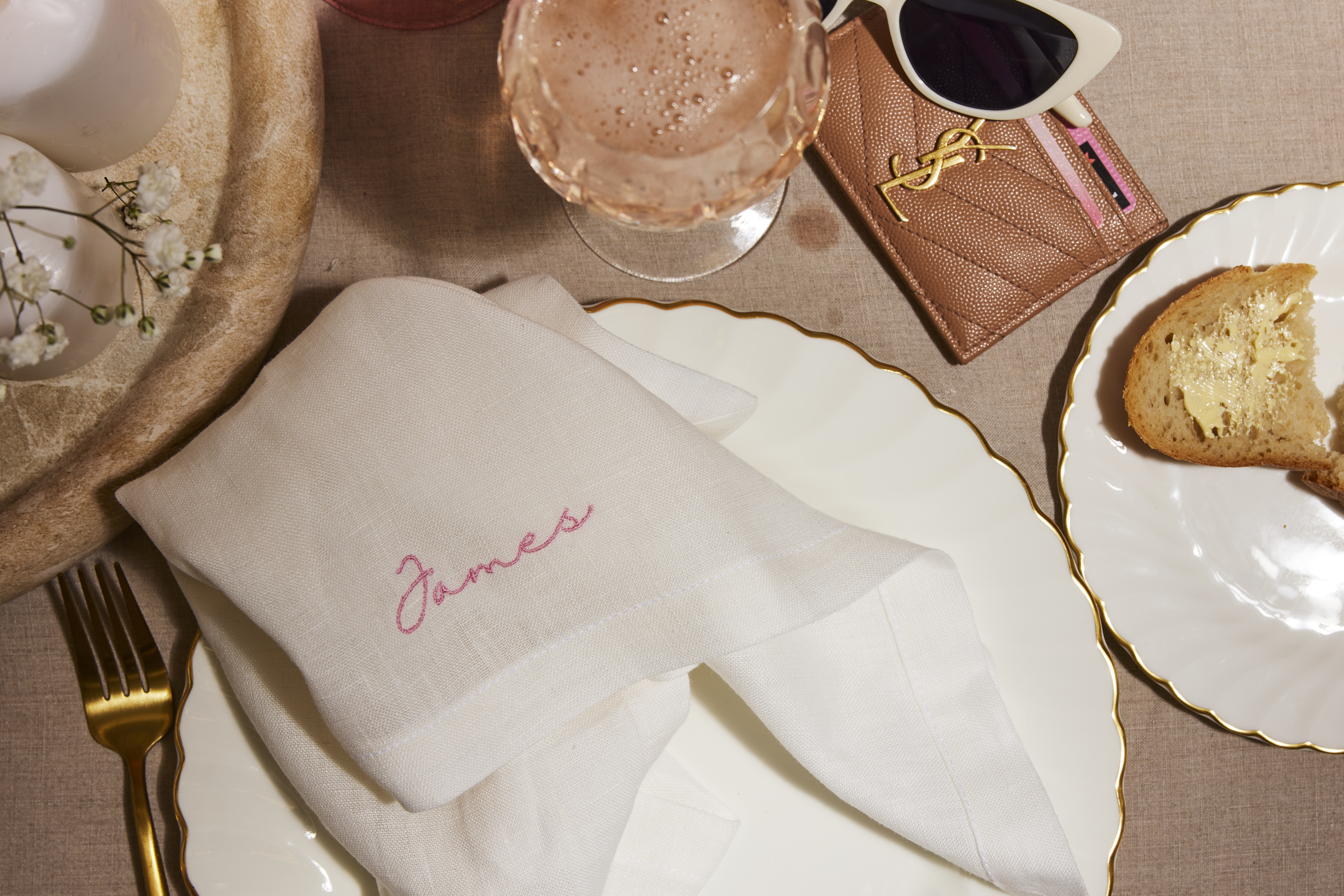 White linen napkin embroidered "James" in pink, folded on a scalloped gold-rimmed plate with a gold fork, pink glass and toast