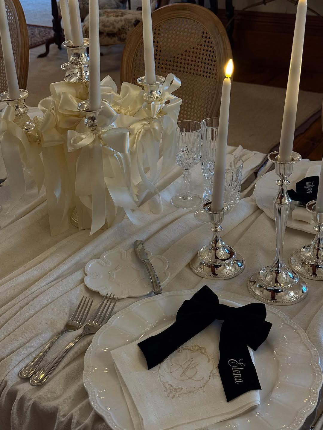 White embroidered linen napkin on a scalloped plate, topped by a black bow reading Elena, with silver candlesticks and cream ribbon‑wrapped tapers