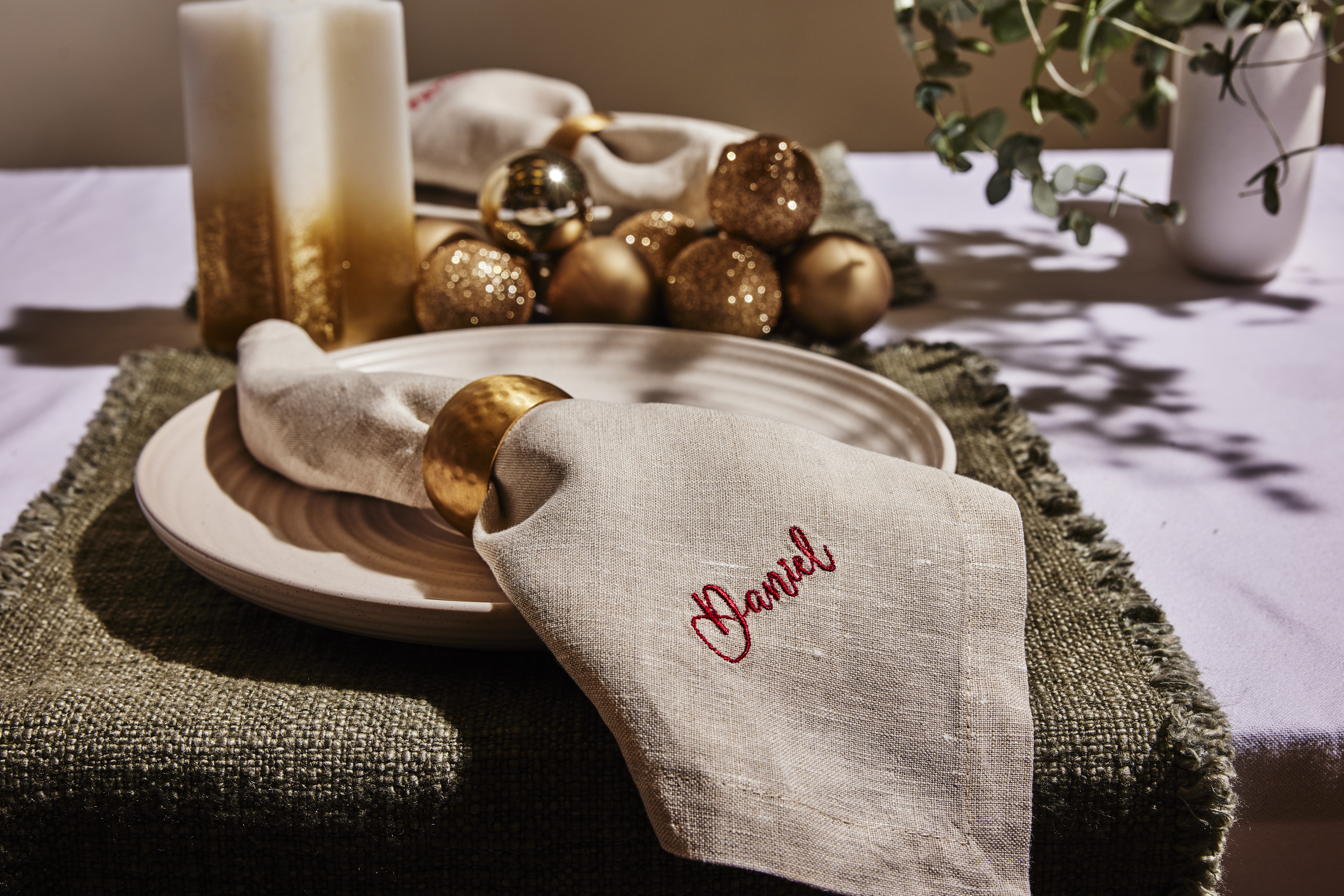Beige linen napkin embroidered "Daniel" in red, gold napkin ring on cream plate over green textured placemat with gold ornaments