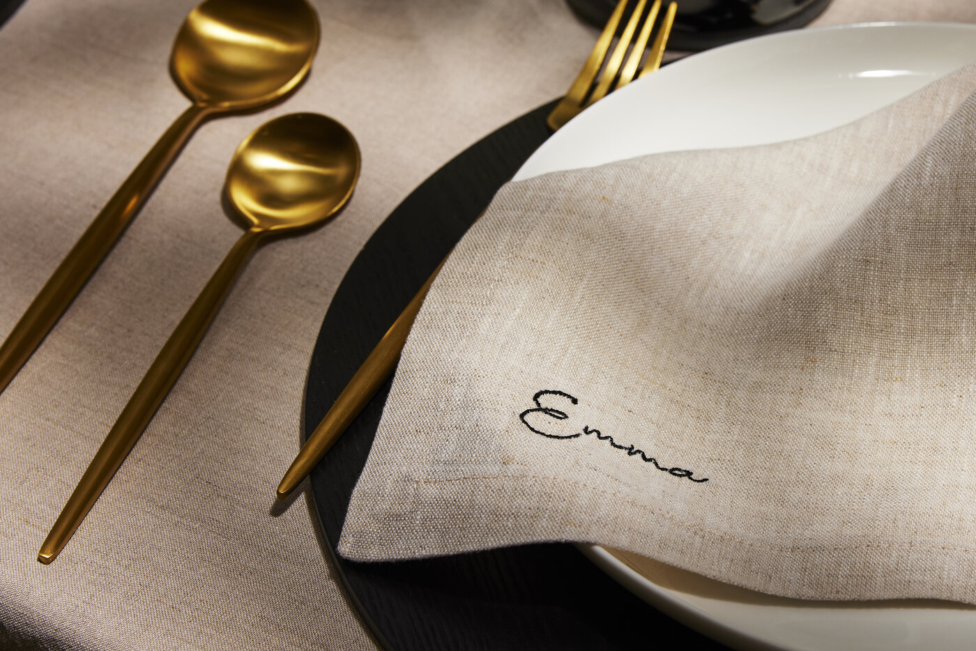 Sand linen napkin embroidered with the name Emma in fine script, styled on a charcoal plate with matte gold cutlery.