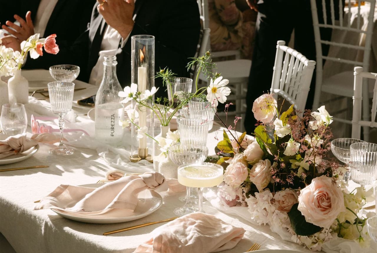 A sunlit wedding tablescape with a blush linen napkin, soft blooms and candlelight
