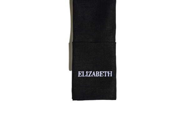 Black linen napkin folded neatly, embroidered with the name Elizabeth in white serif lettering.