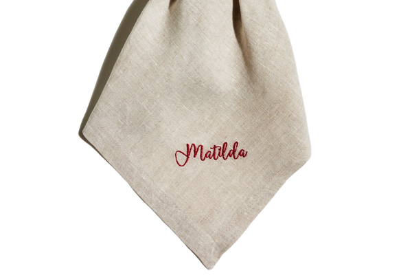Sand linen napkin tied in a knot, embroidered with the name Matilda in red script.